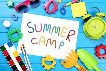 Text Summer camp with plastic toys, sunglasses and paints on blue wooden table