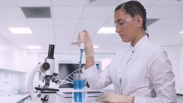 A female lab technician transfers a blue fluid sample to different tubes using a micropipette while sitting at a table next to a laptop and a microscope in a science lab.