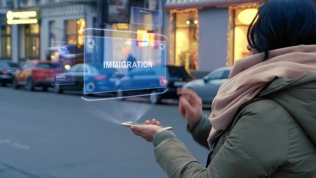 Unrecognizable woman standing on the street interacts HUD hologram with text Immigration. Girl in warm clothes uses technology of the future mobile screen on background of night city