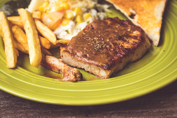 Pig steak is a fast food,pig steak this dish consists of with salad which has  purple lettuce corn apple cabbage salad dressing bread french fries and black pepper sauce, all in a green plate.