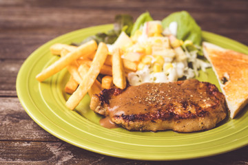 Pig steak is a fast food,pig steak this dish consists of with salad which has  purple lettuce corn apple cabbage salad dressing bread french fries and black pepper sauce, all in a green plate.
