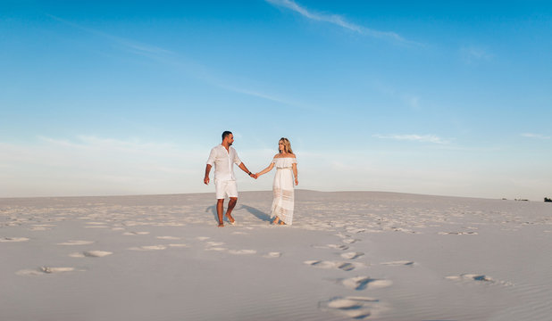 Young beautiful couple of a blonde woman and a sunburned guy hold hands.Happy romantic lovers walk barefoot on the sand in white clothes at sunset. love, honeymoon, and idyllic relationships.Blue sky.