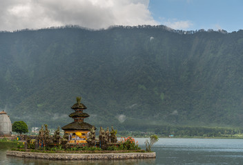 Bali, Indonesia - February 25, 2019: Ulun Danu Beratan Temple complex in Bedoegoel. Linga Petak pagoda of three levels on island under rainy cloudscape. Green and flowers.