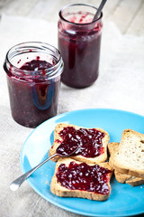 Fresh cereal bread slices on blue ceramic plate, homemade cherry and wild berries jam in jars, ...