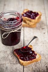 Toasted cereal bread slices and jar with homemade wild berries jam and spoon closeup on rustic wooden table background.