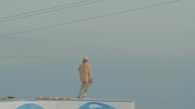 Teenage boy standing on top of a skate park bowl overlooking the ocean in Morocco at sunset with hazy skies