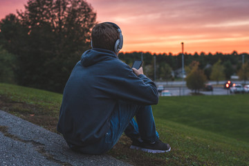Teenager sitting alone at the top of a hill at sunset. He is listening to music through his...