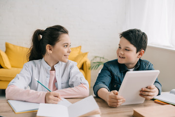 smiling boy using digital tablet while sitting at desk near sister writing in notebook © LIGHTFIELD STUDIOS