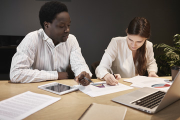 Beautiful young woman having business meeting with dark skinned CEO at his office desk. Black male boss pointing at contract, giving explanations to new female employee, showing her where to sign