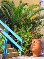 Fototapeta na wymiar Picturesque view on the island of Crete - stairs leading to the house, surrounded by green palm tree and red flowers. Large ceramic pitcher standing by the stairs with blue railings. Crete, Greece.
