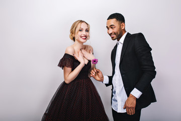 Happy cute couple in-love celebrating Valentine s day on white background. Attractive young blonde woman in luxury dress, handsome man in tuxedo, giving flower, smiling, positive emotions