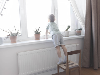 The little boy is standing on the children's chair and looking out the window