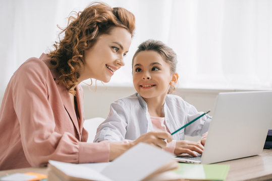 happy mother pointing at laptop screen while helping daughter doing homework