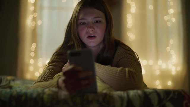 Girl laying on bed at night reading disturbing social media on cell phone / Cedar Hills, Utah, United States