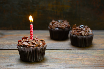 chocolate cupcakes with a burning candle on rustic wood, dark brown background with copy space