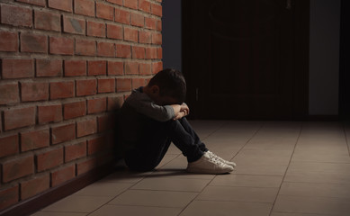 Sad little boy sitting on floor near brick wall indoors