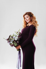 Portrait of young pregnant woman with big flower in her hand. Beautiful model posing in dark dress and holding her stomach at white studio background. Beautiful and happy pregnancy concept