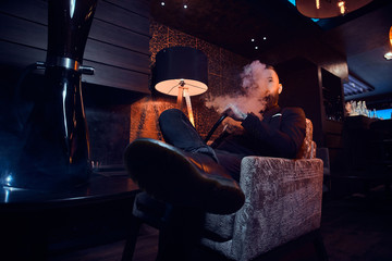 Pensive tattooed man is relaxing on the armchair and is smoking hookah, making hazy vapour.