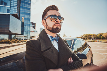 Smart serious man in coat is standing near his new car.