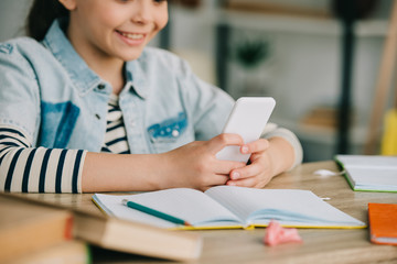 partial view of smiling child using smartphone while sitting at desk and doing schoolwork at home