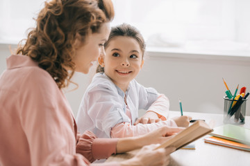 cheerful child looking at beautiful mother while doing homework together - obrazy, fototapety, plakaty
