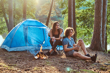 Couple of students are relaxing after long hike in sunny forest.They have tent and bonfire. © Fxquadro