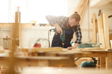 Waist up portrait of red haired carpenter drilling wood while working in joinery lit by sunlight,...