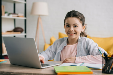 happy child looking at camera while sitting at desk and using laptop at home