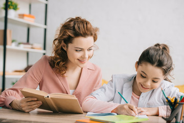 beautiful woman holding book and adorable daughter writing in notebook while doing homework together