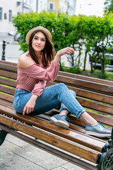 Fashionably dressed young pretty woman on the street on a sunny summer evening. Beautiful girl in jeans, a blouse, small hat and sunglass is sitting on a bench and smiling. Street-fashion concept
