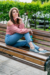 Fashionably dressed young pretty woman on the street on a sunny summer evening. Beautiful girl in jeans, a blouse, small hat and sunglass is sitting on a bench and smiling. Street-fashion concept