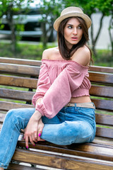 Fashionably dressed young woman on the street on a sunny evening. Beautiful girl in jeans, blouse and small hat is sitting on a bench. Street-fashion concept. Late spring and early summer