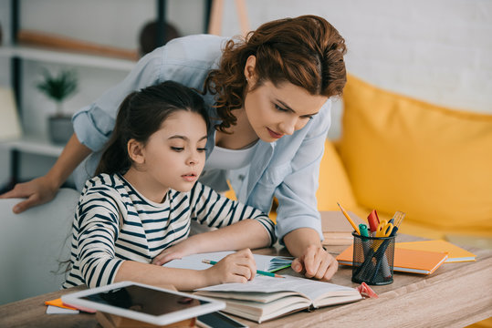 attentive mother helping adorable daughter doing schoolwork at home