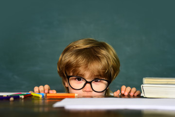 Cute boy with happy face expression near desk with school supplies. Kid is learning in class on background of blackboard.