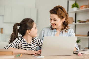 happy mother looking at adorable daughter while using laptop together