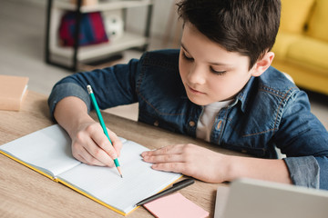 cute attentive boy writing in notebook while sitting at desk and doing homework