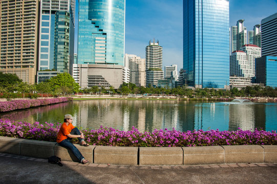tourist girl in bright shirt sitting near blue lake in park in Bangkok, traveler resting near big lake and watching for reflections of skyscrapers