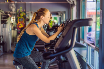 Fototapeta na wymiar Young woman using phone while training at the gym. Woman sitting on exercising machine holding mobile phone