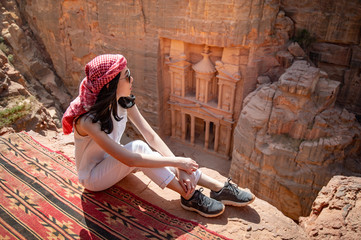 Asian woman traveler sitting on carpet viewpoint in Petra ancient city looking at the Treasury or Al-khazneh, famous travel destination of Jordan and one of seven wonders. UNESCO World Heritage site. - obrazy, fototapety, plakaty