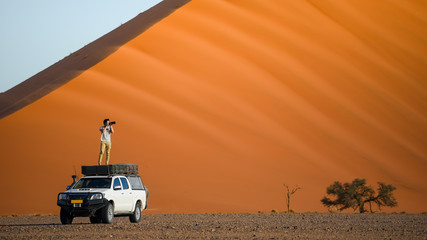 Naklejka na ściany i meble Young Asian man traveler and photographer standing on camper car near orange sand dune. Desert road trip travel concept