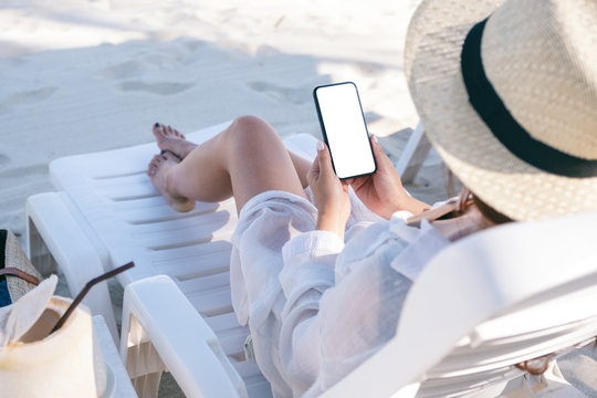Mockup image of a woman holding black mobile phone with blank desktop screen while laying down on beach chair on the beach