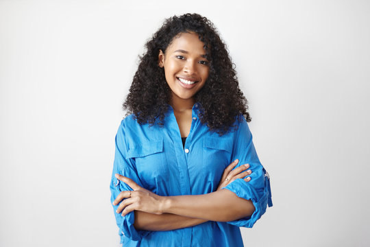 People, lifestyle and ethnicity concept. Portrait of gorgeous positive young dark skinned woman with loose curly hairdo posing isolated in studio, smiling joyfully, crossing arms on her chest