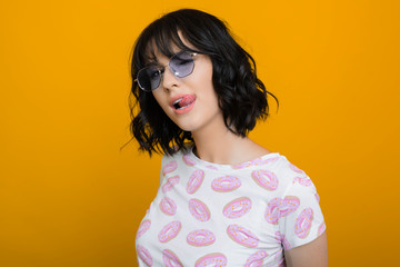 Portrait of a beautiful girl with short dark hair wearing glasses looking at camera flirting while showing tongue against a yellow background.