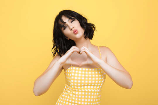 Beautiful stylish caucasian female showing heart symbol while kissing and looking at camera dressed in yellow dress isolated over yellow background.