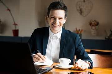 Portrait of a positive adult caucasian manager sitting at his working place while drinking a coffee and looking at camera smiling.