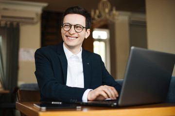 Portrait of a attractive adult freelancer wearing suit and glasses looking away smiling while working at his laptop sitting in a coffee shop.