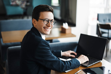 Side view portrait of a stylish adult caucasian manager wearing suit and glasses looking at camera smiling over his shoulder while working at laptop in cafe.