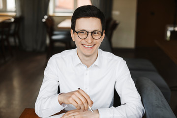 Portrait of a attractive positive manager wearing white shirt and glasses looking away smiling sitting at a desk in a coffee shop waking notes.
