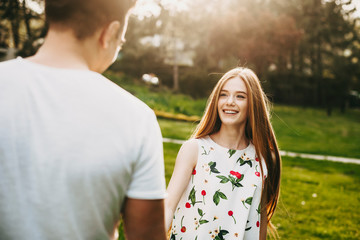 Portrait of lovely caucasian woman with long red hair and freckles looking at his boyfriend laughing against sunset light while holding hands while dating.