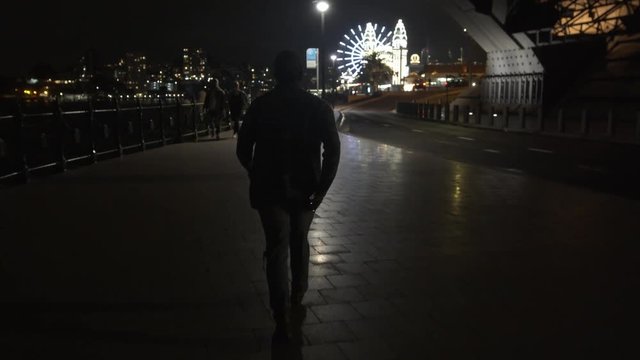 Camera Smoothly Follows Man Walking On Path Towards Theme Park at Night, Wide